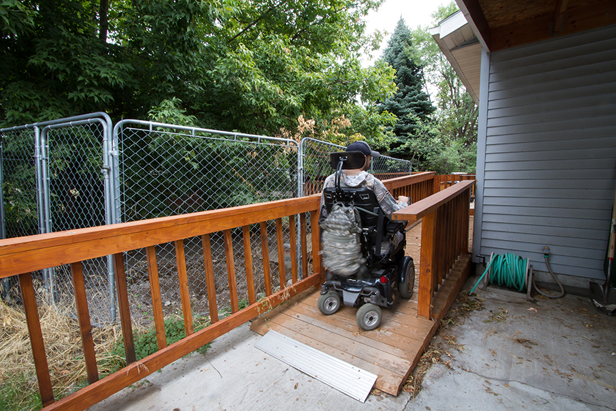 A man wearing a baseball cap uses a power wheelchair to travel up the back wooden ramp toward his rear deck.